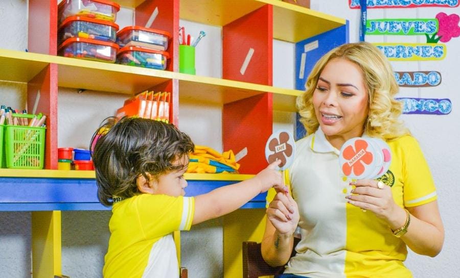 Profesora muestra figuras de colores a un alumno frente al estante de materiales